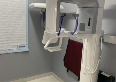 Panoramic dental X-ray machine in a clean imaging room with light-colored flooring and a lead apron hanging nearby