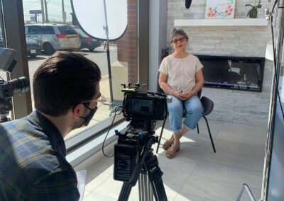 Woman seated near the front window at Avatar Dental during a video interview setup, with lighting, microphone, and a videographer filming the session