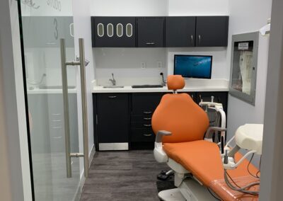 Private dental operatory at Avatar Dental featuring an orange dental chair, overhead light, wall-mounted monitor, and glass entry door